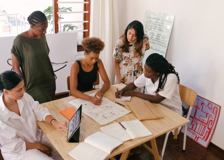 five people of various genders and races/ethnicities are pictured working collaboratively around a table with papers, drawings, tablets on the table